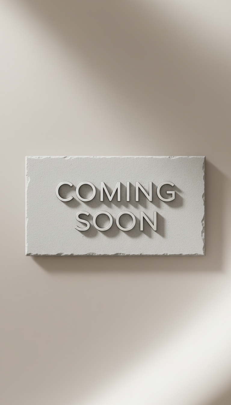 A minimalist, elegantly designed 'coming soon' sign rendered in fine, embossed silver lettering on a textured, pale grey stone plaque. The plaque rests on a soft, muted taupe backdrop featuring gentle, flowing gradients. Subdued, natural side lighting emphasizes the relief of the embossed letters while creating tasteful, diffused shadows for added depth. The visual mood is serene, anticipatory, and distinctly sophisticated. The composition is centered, balanced, and employs a sharp focus, portraying photographic realism and a subtle sense of understated luxury—perfectly communicating the site’s temporary, ‘under construction’ status with elegance.